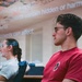 A group of students or attendees are seated in a classroom or lecture setting, attentively watching a presentation. A projected slide is visible overhead discussing dietary supplements, their potential risks, and their effects on health. The foreground subject is a young male wearing glasses and a red t-shirt.