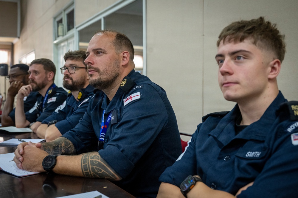 A group of Royal Navy personnel are seated at a table, appearing to attend a briefing or meeting. The individuals are wearing dark navy blue uniforms with visible Royal Navy insignia and name tags. The setting appears to be an indoor conference or briefing room.