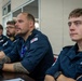 A group of Royal Navy personnel are seated at a table, appearing to attend a briefing or meeting. The individuals are wearing dark navy blue uniforms with visible Royal Navy insignia and name tags. The setting appears to be an indoor conference or briefing room.