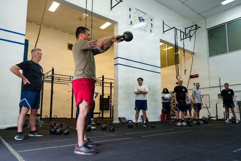A group of individuals are participating in a CrossFit-style workout session inside a gymnasium. One person in the foreground is performing a kettlebell swing while others observe. The setting appears to be a military or institutional fitness facility based on the signage and attire.