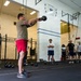 A group of individuals are participating in a CrossFit-style workout session inside a gymnasium. One person in the foreground is performing a kettlebell swing while others observe. The setting appears to be a military or institutional fitness facility based on the signage and attire.