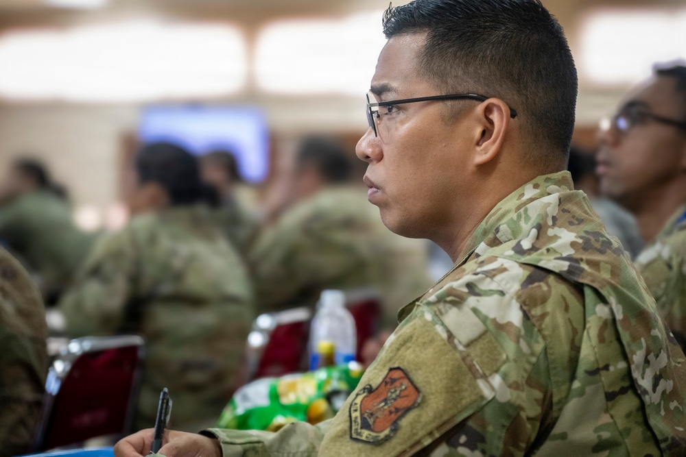 A military service member wearing Operational Camouflage Pattern (OCP) uniform is seated in what appears to be a briefing or training session. The individual is attentively focused forward, holding a pen as if taking notes. Multiple other uniformed personnel are visible in the blurred background, suggesting a group military event or classroom setting.