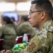 A military service member wearing Operational Camouflage Pattern (OCP) uniform is seated in what appears to be a briefing or training session. The individual is attentively focused forward, holding a pen as if taking notes. Multiple other uniformed personnel are visible in the blurred background, suggesting a group military event or classroom setting.
