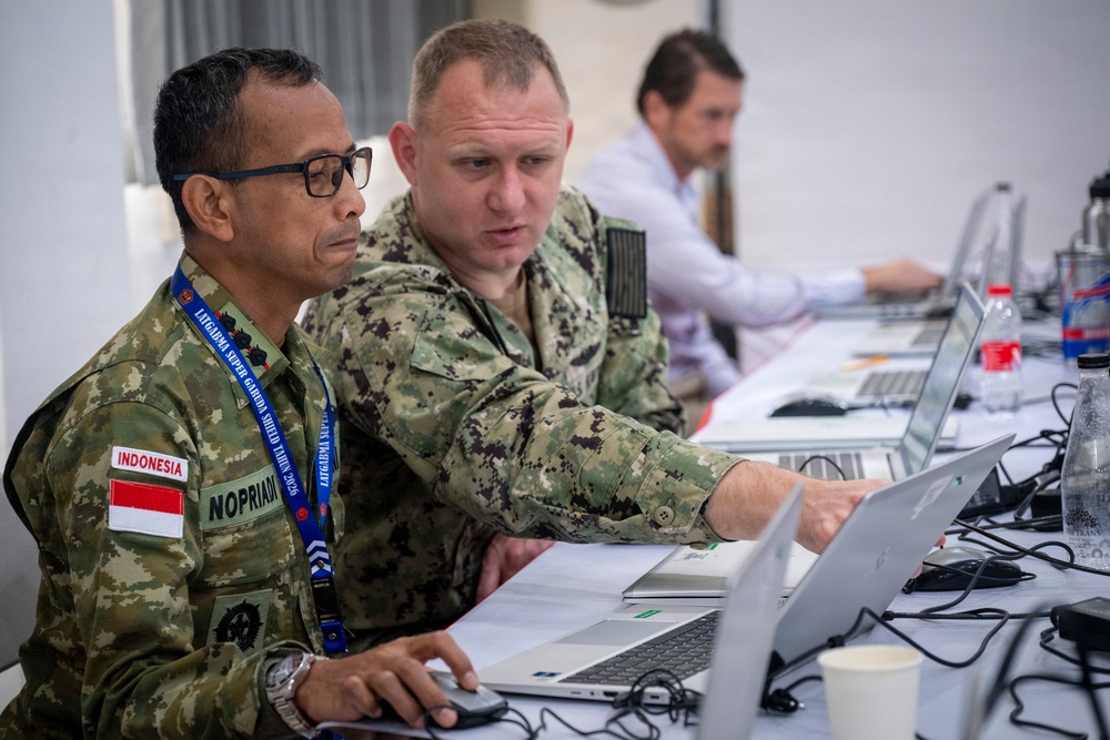 Two military personnel in camouflage uniforms are collaborating at a workstation with laptops in what appears to be a joint military exercise or operations center. The individual on the left wears an Indonesian military uniform with a name tag reading 'NOPRIAD' and a lanyard badge, while the person on the right points at the laptop screen. A third person in civilian attire works in the background.