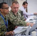 Two military personnel in camouflage uniforms are collaborating at a workstation with laptops in what appears to be a joint military exercise or operations center. The individual on the left wears an Indonesian military uniform with a name tag reading 'NOPRIAD' and a lanyard badge, while the person on the right points at the laptop screen. A third person in civilian attire works in the background.