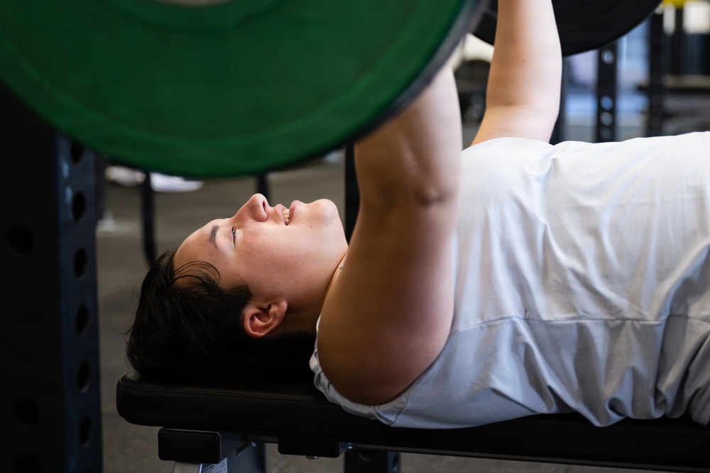 A person is performing a bench press exercise in a gym setting, lying on a flat bench and pressing a barbell loaded with green weight plates upward. The individual is wearing a white t-shirt and appears to be mid-lift with arms extended. The image is taken from a close, side-angle perspective that emphasizes the lifter's face and upper body.