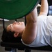 A person is performing a bench press exercise in a gym setting, lying on a flat bench and pressing a barbell loaded with green weight plates upward. The individual is wearing a white t-shirt and appears to be mid-lift with arms extended. The image is taken from a close, side-angle perspective that emphasizes the lifter's face and upper body.