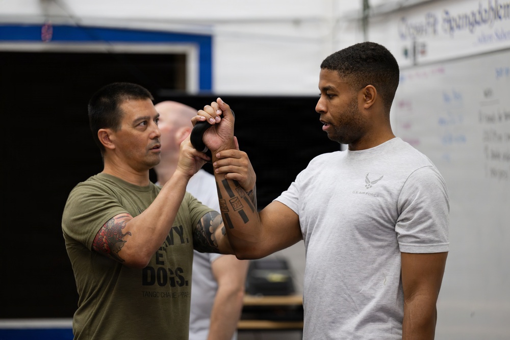 Two men are engaged in what appears to be a kettlebell training or instruction session in a gym or CrossFit-style facility. The man on the left, wearing an olive green t-shirt, is guiding the grip or technique of the man on the right, who is wearing a U.S. Air Force t-shirt. Both individuals have visible tattoos, and a third person is partially visible in the background.