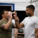 Two men are engaged in what appears to be a kettlebell training or instruction session in a gym or CrossFit-style facility. The man on the left, wearing an olive green t-shirt, is guiding the grip or technique of the man on the right, who is wearing a U.S. Air Force t-shirt. Both individuals have visible tattoos, and a third person is partially visible in the background.