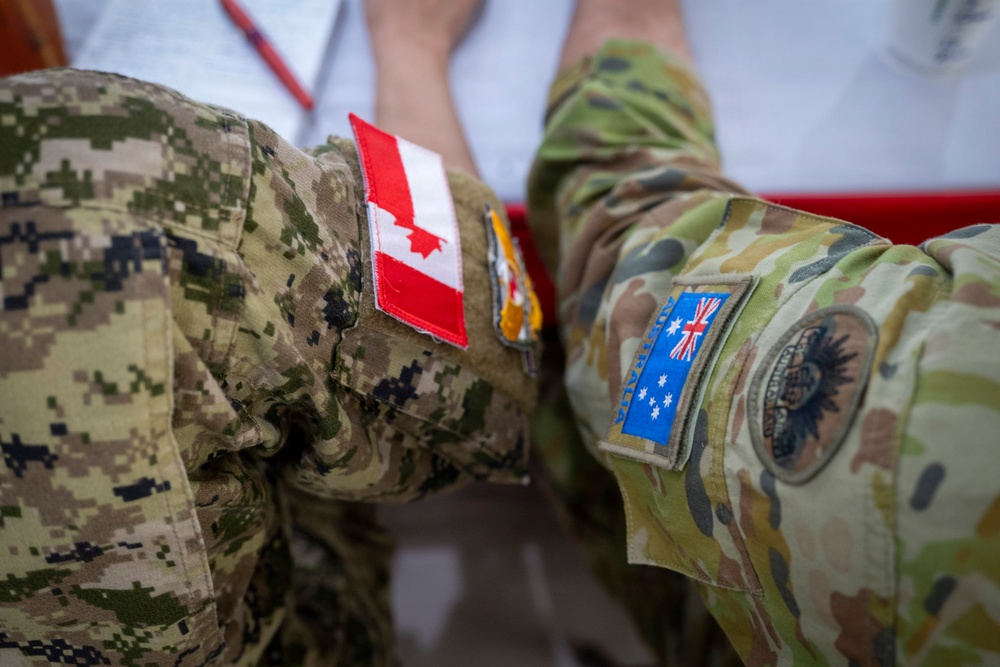 The image shows a close-up of military uniforms worn by personnel from at least two different nations. A Canadian flag patch is visible on a MultiCam/CADPAT-style uniform in the center, while an Australian flag patch and what appears to be a unit insignia are visible on an AUSCAM (Australian Multicam Camouflage Uniform) on the right. A third uniform in a digital camouflage pattern, consistent with US Navy NWU or similar, is visible on the left.