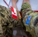 The image shows a close-up of military uniforms worn by personnel from at least two different nations. A Canadian flag patch is visible on a MultiCam/CADPAT-style uniform in the center, while an Australian flag patch and what appears to be a unit insignia are visible on an AUSCAM (Australian Multicam Camouflage Uniform) on the right. A third uniform in a digital camouflage pattern, consistent with US Navy NWU or similar, is visible on the left.