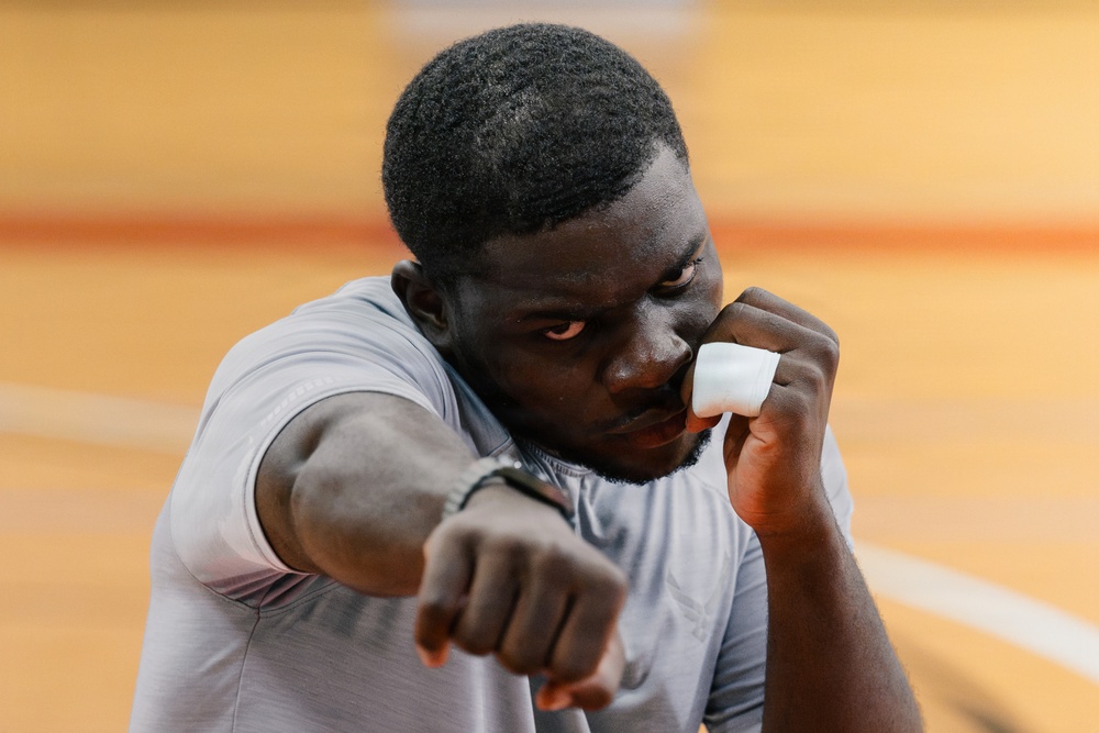 A young man is photographed in a boxing or fighting stance, with one fist extended toward the camera and the other raised near his face in a guard position. He is wearing a gray athletic shirt and has a white finger wrap or tape on one hand. The background appears to be an indoor sports court with an orange/wood-toned floor.