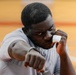 A young man is photographed in a boxing or fighting stance, with one fist extended toward the camera and the other raised near his face in a guard position. He is wearing a gray athletic shirt and has a white finger wrap or tape on one hand. The background appears to be an indoor sports court with an orange/wood-toned floor.