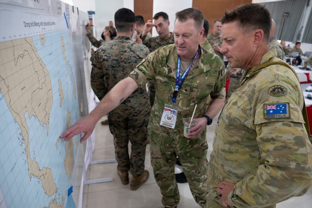 Military personnel from multiple nations are gathered around a large map board during what appears to be a joint military planning or briefing session. At least one soldier is clearly identified as Australian by the flag patch on their uniform, while others appear to be US Marines based on their distinctive MARPAT camouflage. The setting appears to be an indoor conference or operations room with tables and equipment visible in the background.