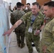 Military personnel from multiple nations are gathered around a large map board during what appears to be a joint military planning or briefing session. At least one soldier is clearly identified as Australian by the flag patch on their uniform, while others appear to be US Marines based on their distinctive MARPAT camouflage. The setting appears to be an indoor conference or operations room with tables and equipment visible in the background.