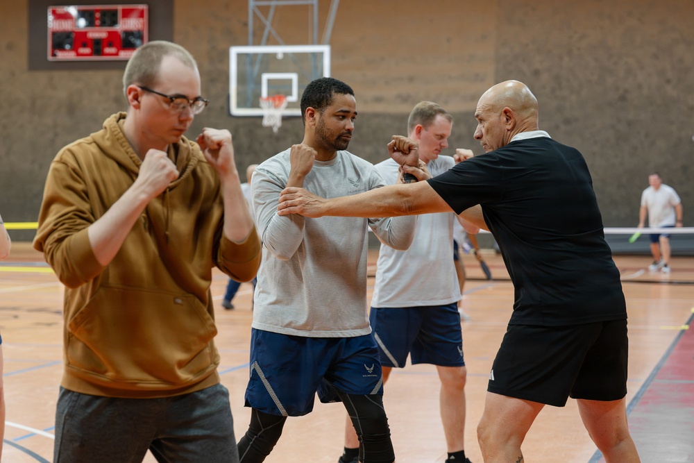 A group of individuals are participating in a self-defense or martial arts training session inside a gymnasium. A bald instructor in a black shirt appears to be demonstrating techniques with a student in a gray shirt, while others practice nearby. The setting is an indoor sports facility with a basketball hoop visible in the background.