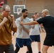 A group of individuals are participating in a self-defense or martial arts training session inside a gymnasium. A bald instructor in a black shirt appears to be demonstrating techniques with a student in a gray shirt, while others practice nearby. The setting is an indoor sports facility with a basketball hoop visible in the background.