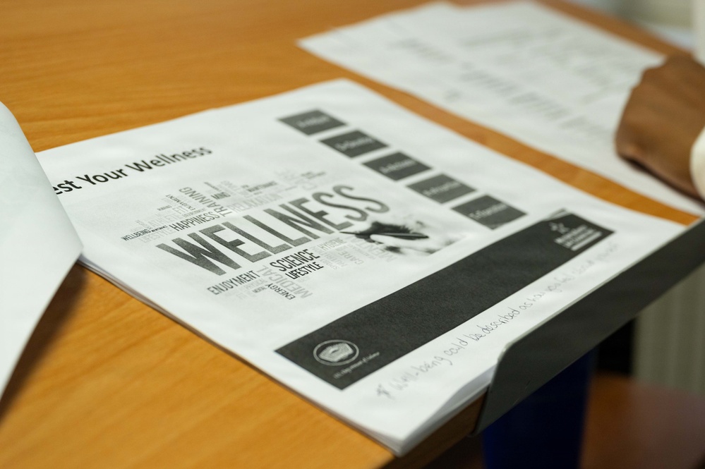 The image shows printed wellness-related documents laid open on a wooden desk. The main visible document features a word cloud design centered around the word 'WELLNESS' along with related terms. Additional papers are visible in the background, and handwritten notes appear on one of the documents.