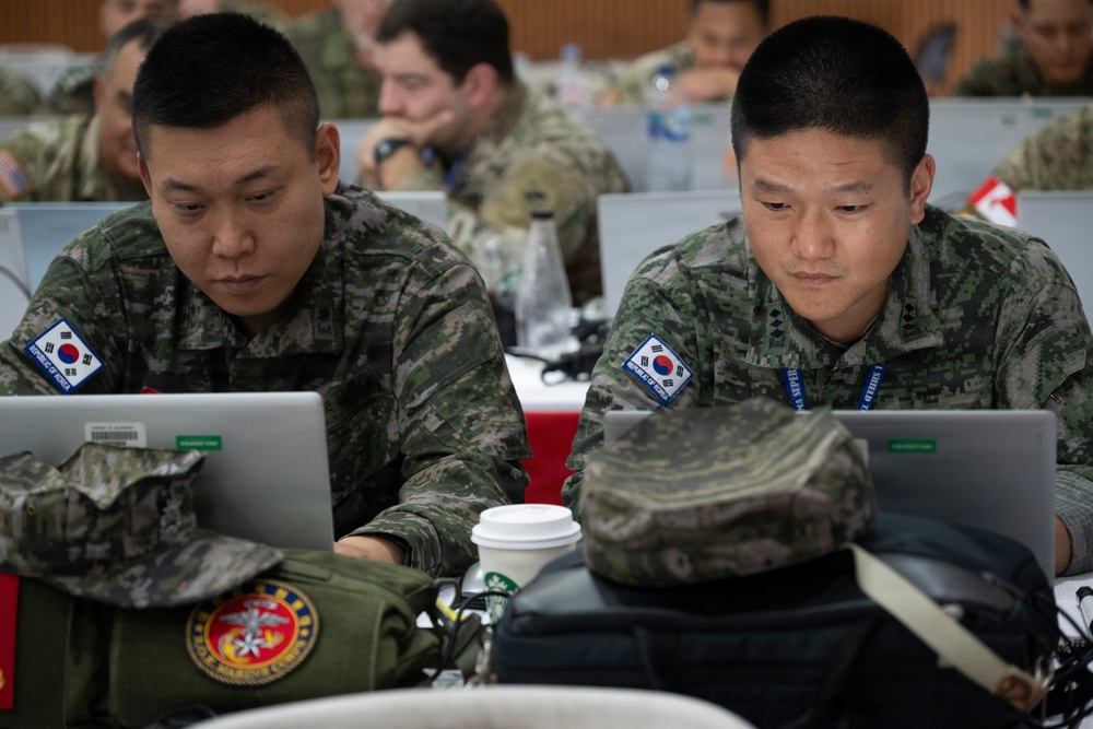 Two Republic of Korea (ROK) military personnel are working at laptops in what appears to be a multinational military briefing or exercise environment. Both soldiers wear South Korean digital camouflage uniforms with South Korean flag patches visible on their shoulders. The setting suggests a joint military exercise or conference with multiple nations represented in the background.