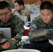 Two Republic of Korea (ROK) military personnel are working at laptops in what appears to be a multinational military briefing or exercise environment. Both soldiers wear South Korean digital camouflage uniforms with South Korean flag patches visible on their shoulders. The setting suggests a joint military exercise or conference with multiple nations represented in the background.
