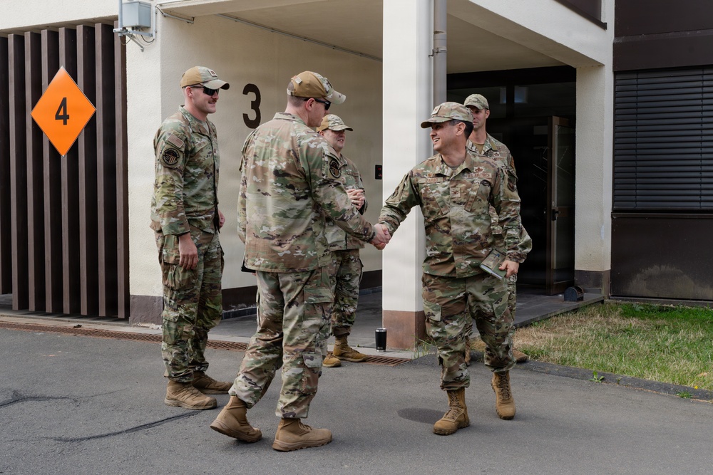 Four military personnel in OCP (Operational Camouflage Pattern) uniforms are gathered outside a building marked with the number '3' and an orange diamond hazard sign marked '4'. Two of the individuals are shaking hands in what appears to be a greeting or formal meeting. The setting appears to be a military installation or base facility.