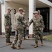 Four military personnel in OCP (Operational Camouflage Pattern) uniforms are gathered outside a building marked with the number '3' and an orange diamond hazard sign marked '4'. Two of the individuals are shaking hands in what appears to be a greeting or formal meeting. The setting appears to be a military installation or base facility.