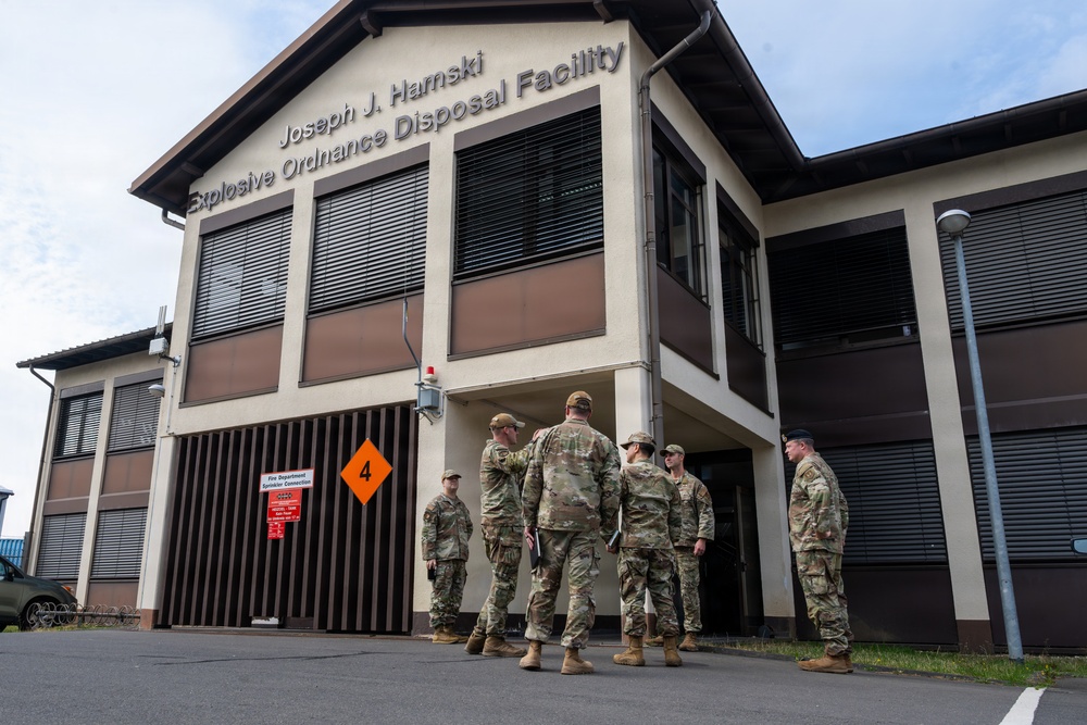 A group of military personnel in camouflage uniforms are gathered outside the Joseph J. Harnski Explosive Ordnance Disposal (EOD) Facility. The two-story building features brown and beige exterior with horizontal shuttered windows and a hazard diamond placard marked with the number 4. The soldiers appear to be in a briefing or discussion near the building's entrance.
