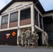 A group of military personnel in camouflage uniforms are gathered outside the Joseph J. Harnski Explosive Ordnance Disposal (EOD) Facility. The two-story building features brown and beige exterior with horizontal shuttered windows and a hazard diamond placard marked with the number 4. The soldiers appear to be in a briefing or discussion near the building's entrance.