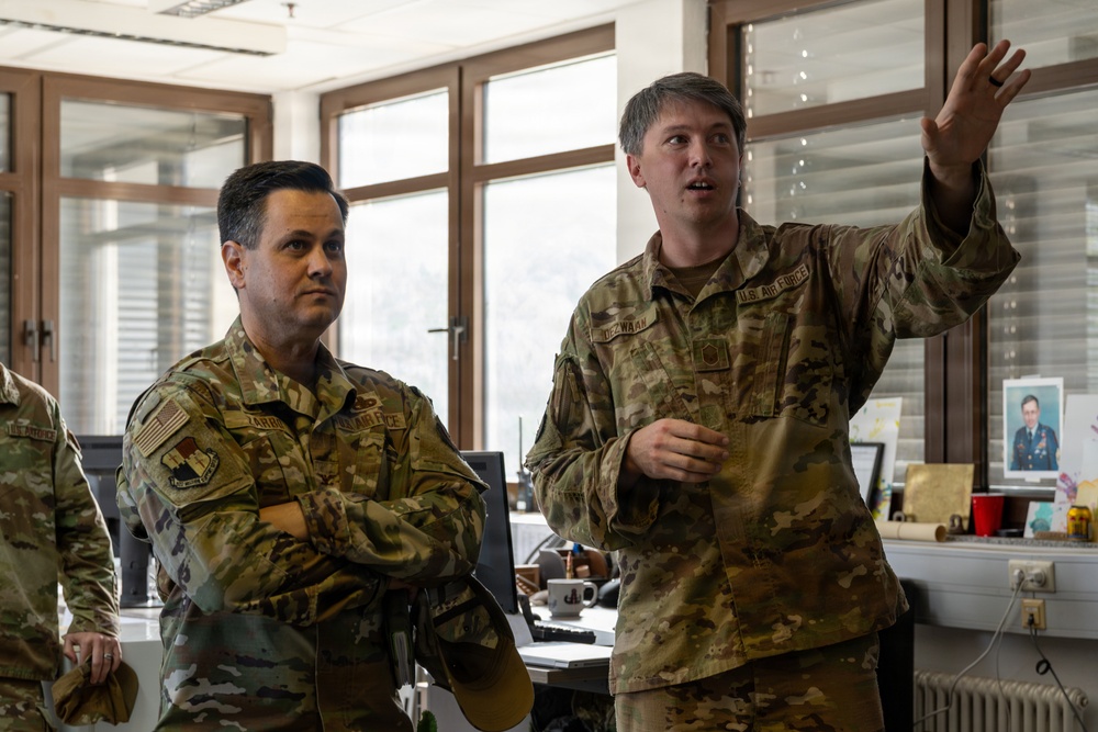 Two U.S. Air Force personnel in OCP (Operational Camouflage Pattern) uniforms are engaged in a briefing or discussion inside what appears to be a military office environment. The individual on the right is gesturing with his raised hand, apparently explaining or pointing something out, while the individual on the left listens attentively with arms crossed. A third partially visible service member is present on the far left.