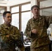 Two U.S. Air Force personnel in OCP (Operational Camouflage Pattern) uniforms are engaged in a briefing or discussion inside what appears to be a military office environment. The individual on the right is gesturing with his raised hand, apparently explaining or pointing something out, while the individual on the left listens attentively with arms crossed. A third partially visible service member is present on the far left.