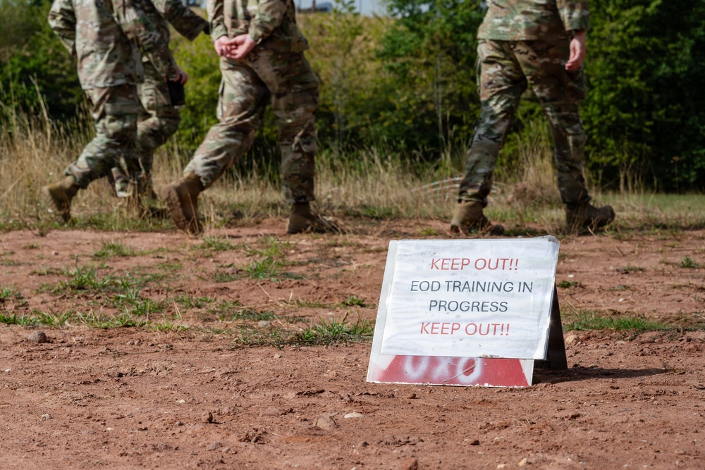 A military training exercise is underway at an outdoor field location. A tent-style warning sign in the foreground reads 'KEEP OUT!! EOD TRAINING IN PROGRESS KEEP OUT!!' in red and black text. Multiple soldiers in camouflage uniforms and combat boots are walking in the background on red dirt terrain.