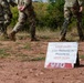 A military training exercise is underway at an outdoor field location. A tent-style warning sign in the foreground reads 'KEEP OUT!! EOD TRAINING IN PROGRESS KEEP OUT!!' in red and black text. Multiple soldiers in camouflage uniforms and combat boots are walking in the background on red dirt terrain.