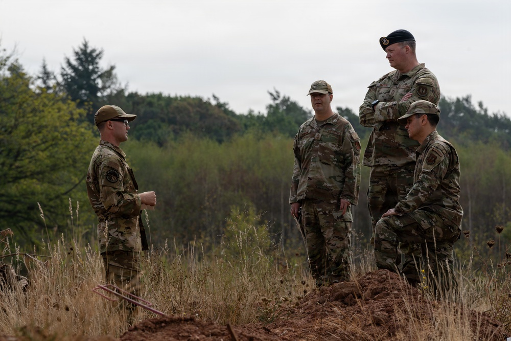 Four military personnel in camouflage uniforms are gathered outdoors in a field or training area, engaged in what appears to be a briefing or discussion. One individual wearing a black beret stands elevated on a mound of disturbed earth, suggesting a position of authority or observation. The setting appears to be a wooded or semi-rural training environment with overcast skies.