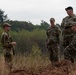 Four military personnel in camouflage uniforms are gathered outdoors in a field or training area, engaged in what appears to be a briefing or discussion. One individual wearing a black beret stands elevated on a mound of disturbed earth, suggesting a position of authority or observation. The setting appears to be a wooded or semi-rural training environment with overcast skies.