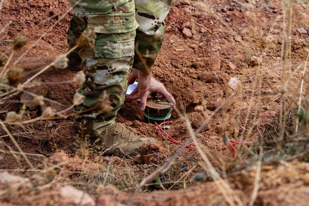 A person wearing military-pattern camouflage clothing and tactical boots is crouching near a partially excavated object in red-brown soil. The object appears to be a landmine or IED (improvised explosive device) with visible red wires extending from it. The scene suggests EOD (Explosive Ordnance Disposal) training or a demining operation.