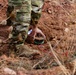 A person wearing military-pattern camouflage clothing and tactical boots is crouching near a partially excavated object in red-brown soil. The object appears to be a landmine or IED (improvised explosive device) with visible red wires extending from it. The scene suggests EOD (Explosive Ordnance Disposal) training or a demining operation.