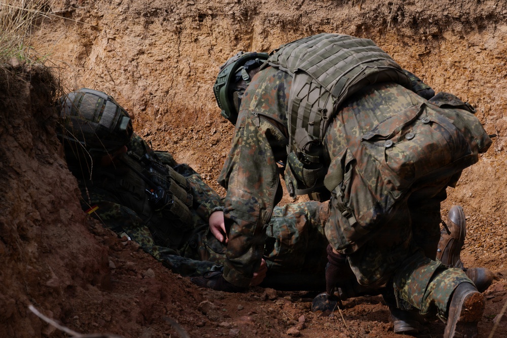 Two soldiers in Flecktarn (German Bundeswehr) camouflage uniforms are positioned inside a dirt trench or foxhole. One soldier appears to be assisting or checking on the other who is lower in the trench. The setting suggests a field training exercise based on the freshly excavated earth and tactical gear worn.