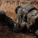 Two soldiers in Flecktarn (German Bundeswehr) camouflage uniforms are positioned inside a dirt trench or foxhole. One soldier appears to be assisting or checking on the other who is lower in the trench. The setting suggests a field training exercise based on the freshly excavated earth and tactical gear worn.