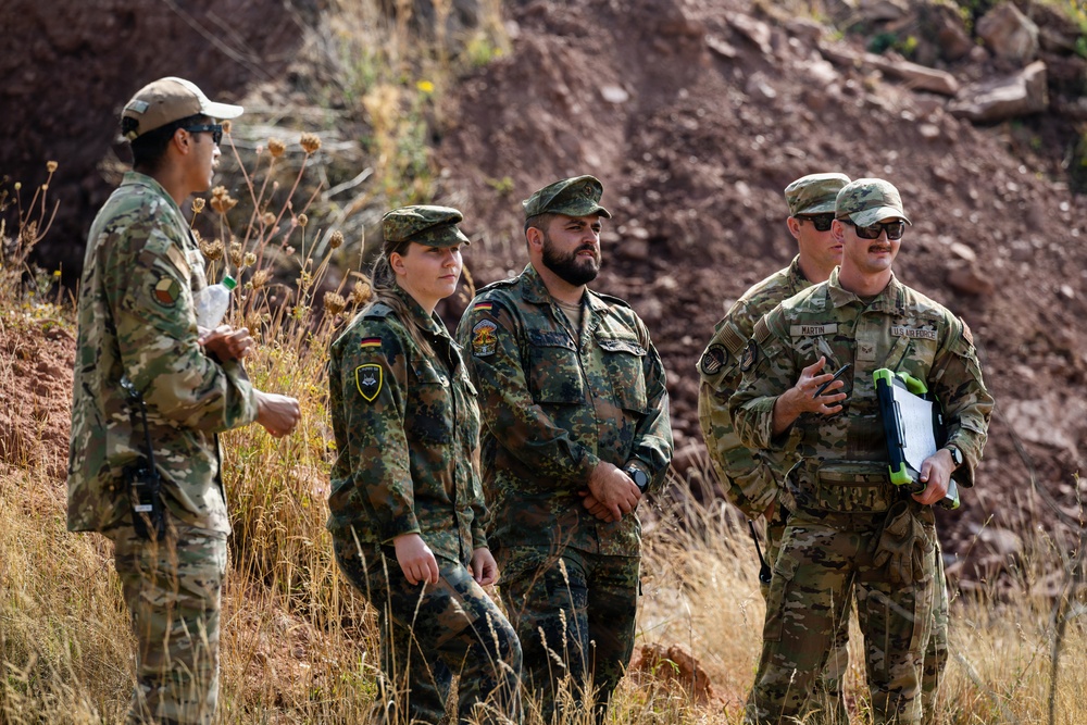 The image shows a group of five military personnel standing outdoors in a rocky, arid terrain. Two individuals are wearing German Bundeswehr flecktarn camouflage uniforms with visible German flag patches, while the others wear US military MultiCam/OCP uniforms, including one clearly identified as US Air Force. The group appears to be engaged in a briefing or discussion, suggesting a joint multinational military exercise or training event.