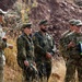 The image shows a group of five military personnel standing outdoors in a rocky, arid terrain. Two individuals are wearing German Bundeswehr flecktarn camouflage uniforms with visible German flag patches, while the others wear US military MultiCam/OCP uniforms, including one clearly identified as US Air Force. The group appears to be engaged in a briefing or discussion, suggesting a joint multinational military exercise or training event.