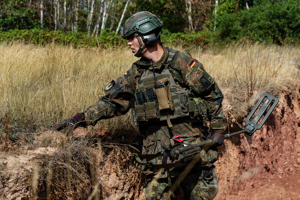 A German military soldier in full combat gear is navigating through a field environment, carrying what appears to be a mine detector or metal detection device. The soldier is wearing Bundeswehr flecktarn camouflage pattern uniform with visible German flag patches on both shoulders. The setting appears to be a training exercise or field operation in a sandy, vegetated terrain.
