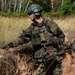 A German military soldier in full combat gear is navigating through a field environment, carrying what appears to be a mine detector or metal detection device. The soldier is wearing Bundeswehr flecktarn camouflage pattern uniform with visible German flag patches on both shoulders. The setting appears to be a training exercise or field operation in a sandy, vegetated terrain.