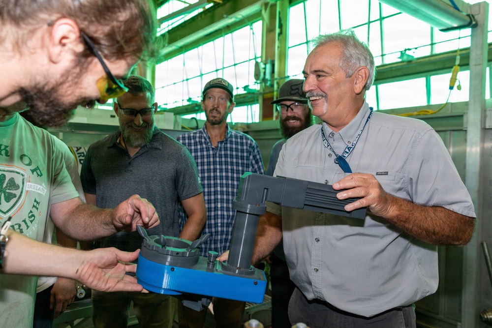 A group of five men are gathered in what appears to be an industrial or workshop facility, examining a blue and gray mechanical component or device. An older man with gray hair and a lanyard is holding and presenting the object while others look on with interest. The setting features large industrial windows with green-tinted lighting overhead.