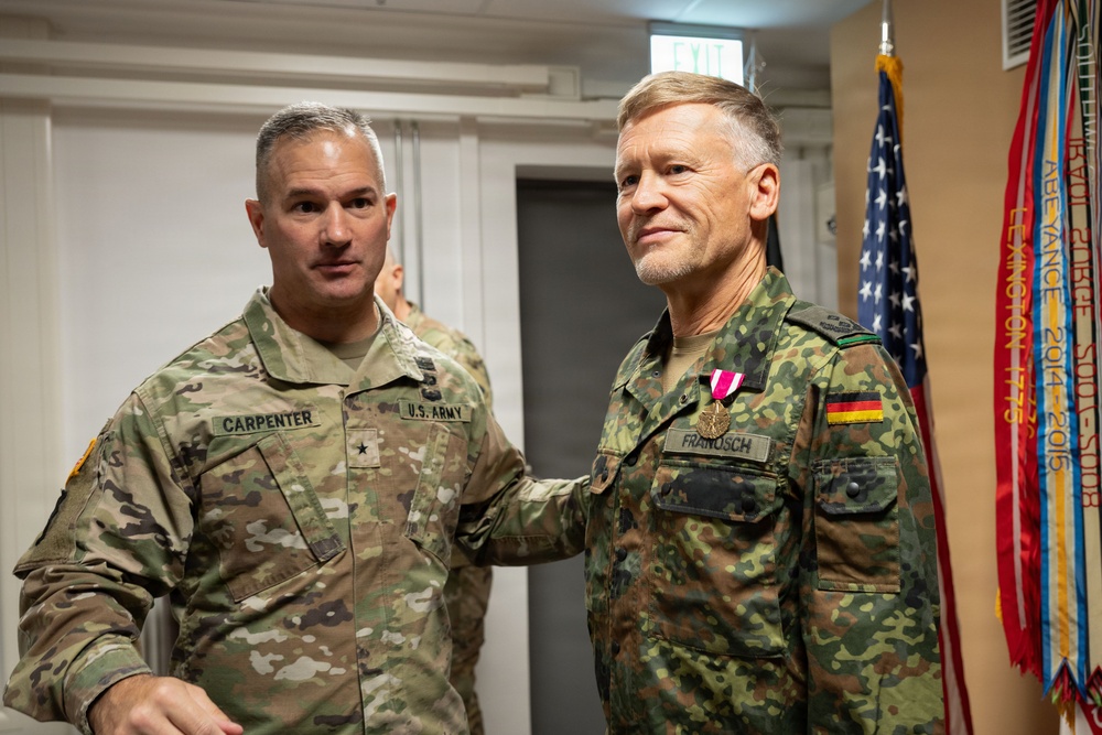 Two military officers are posing together in what appears to be an official ceremony or award presentation. The officer on the left wears a U.S. Army uniform with the name tag 'CARPENTER' and a Brigadier General star insignia, while the officer on the right wears a German Bundeswehr uniform with the name tag 'FRANOSCH' and is displaying a newly awarded medal. An American flag and military unit guidons are visible in the background.