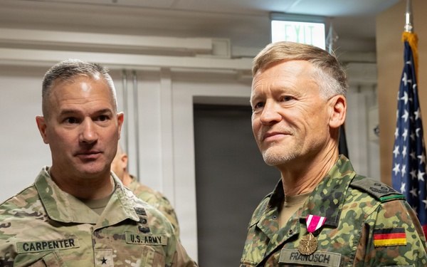 Two military officers are posing together in what appears to be an official ceremony or award presentation. The officer on the left wears a U.S. Army uniform with the name tag 'CARPENTER' and a Brigadier General star insignia, while the officer on the right wears a German Bundeswehr uniform with the name tag 'FRANOSCH' and is displaying a newly awarded medal. An American flag and military unit guidons are visible in the background.