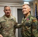 Two military officers are posing together in what appears to be an official ceremony or award presentation. The officer on the left wears a U.S. Army uniform with the name tag 'CARPENTER' and a Brigadier General star insignia, while the officer on the right wears a German Bundeswehr uniform with the name tag 'FRANOSCH' and is displaying a newly awarded medal. An American flag and military unit guidons are visible in the background.