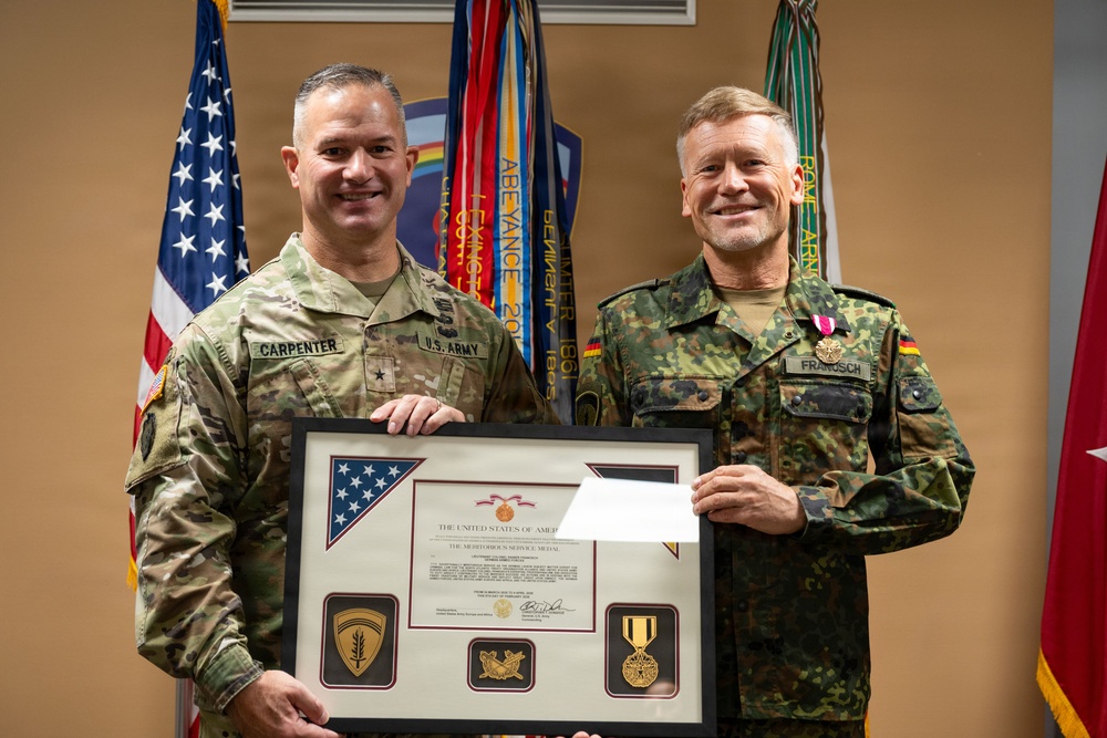 Two military officers pose together holding a framed award certificate. The officer on the left wears a U.S. Army uniform with the name tag 'CARPENTER' and a brigadier general star, while the officer on the right wears a German Bundeswehr uniform with the name tag 'FRANOSCH' and a medal pinned to his chest. The framed document appears to be a Meritorious Service Medal citation from the United States of America.