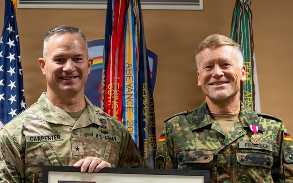 Two military officers pose together holding a framed award certificate. The officer on the left wears a U.S. Army uniform with the name tag 'CARPENTER' and a brigadier general star, while the officer on the right wears a German Bundeswehr uniform with the name tag 'FRANOSCH' and a medal pinned to his chest. The framed document appears to be a Meritorious Service Medal citation from the United States of America.
