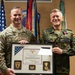 Two military officers pose together holding a framed award certificate. The officer on the left wears a U.S. Army uniform with the name tag 'CARPENTER' and a brigadier general star, while the officer on the right wears a German Bundeswehr uniform with the name tag 'FRANOSCH' and a medal pinned to his chest. The framed document appears to be a Meritorious Service Medal citation from the United States of America.