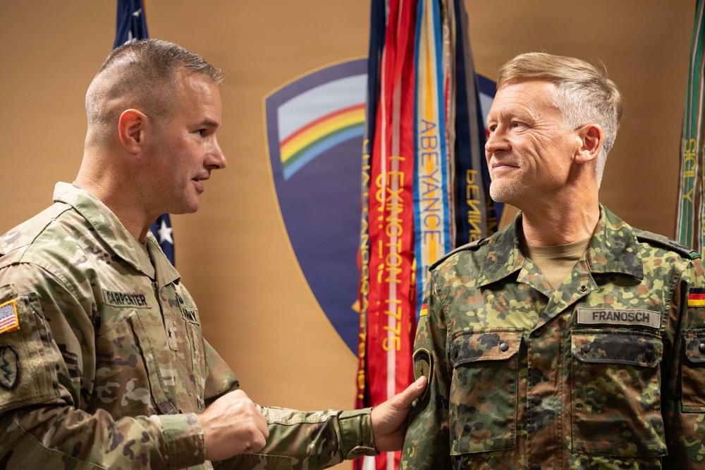 Two military officers are engaged in what appears to be a formal ceremony or award presentation. The officer on the left, identified by his name tape as 'CARPENTER,' is a U.S. Army soldier presenting or pinning something to the officer on the right, identified as 'FRANOSCH,' who is wearing a German Bundeswehr uniform. They are standing in front of military flags and a unit crest backdrop.